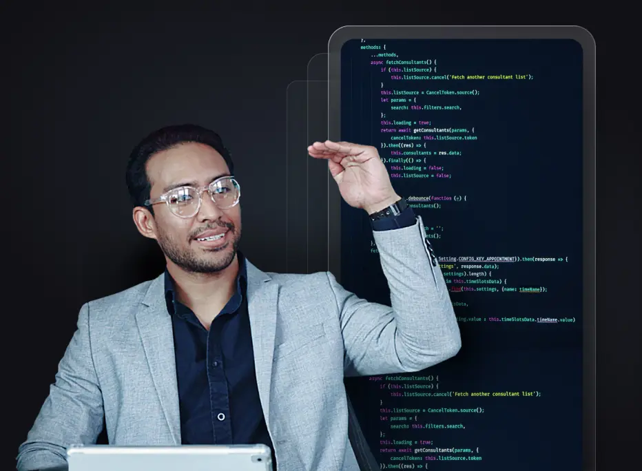 A man wearing glasses and a light gray blazer gestures with one hand raised while sitting in front of a tablet, with large transparent screens behind him displaying colorful lines of computer code.