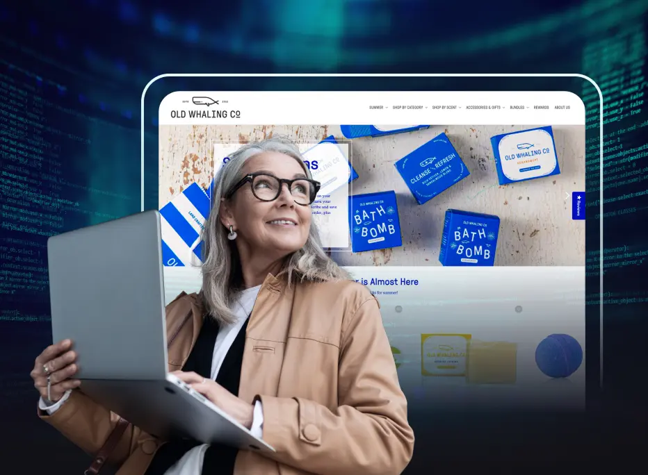 A smiling woman with gray hair and glasses holds an open laptop while looking upward. She stands in front of a large digital display showing a website for ‘Old Whaling Co,’ featuring blue bath bomb products. The background includes faint lines of computer code.