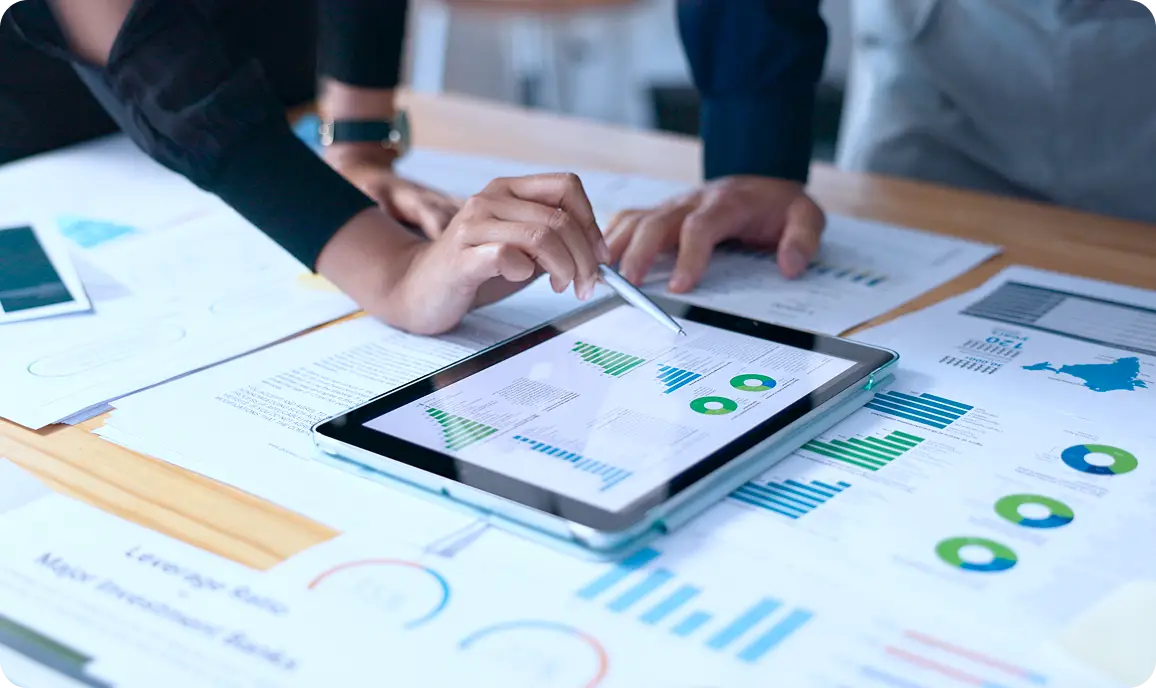 <p>Close-up of two people reviewing data, with one person using a stylus on a tablet displaying charts and graphs, surrounded by printed reports with colorful analytics visuals on a desk.</p>