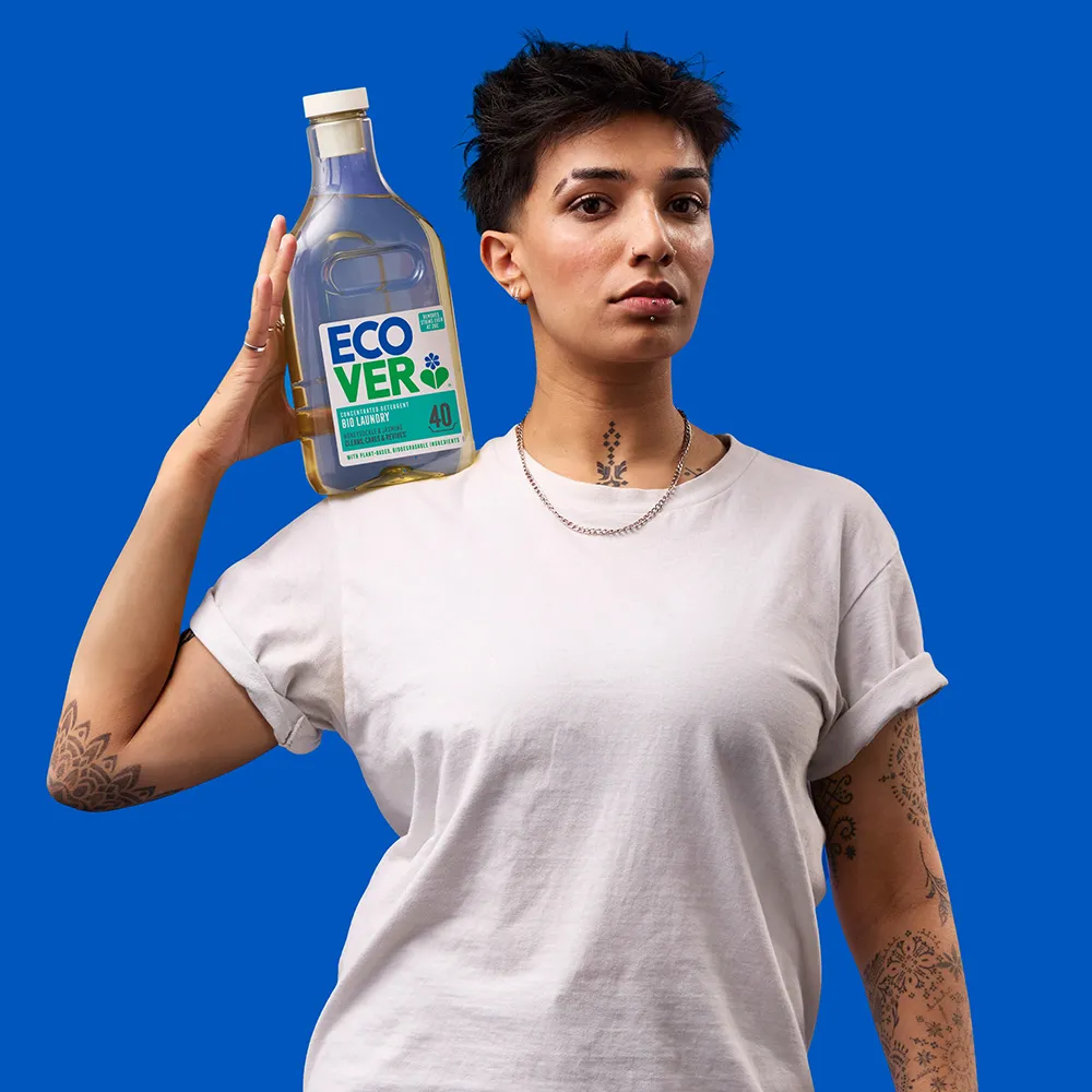 A person with short dark hair wearing a white t-shirt holds a large bottle of Ecover laundry detergent on their shoulder against a solid blue background.