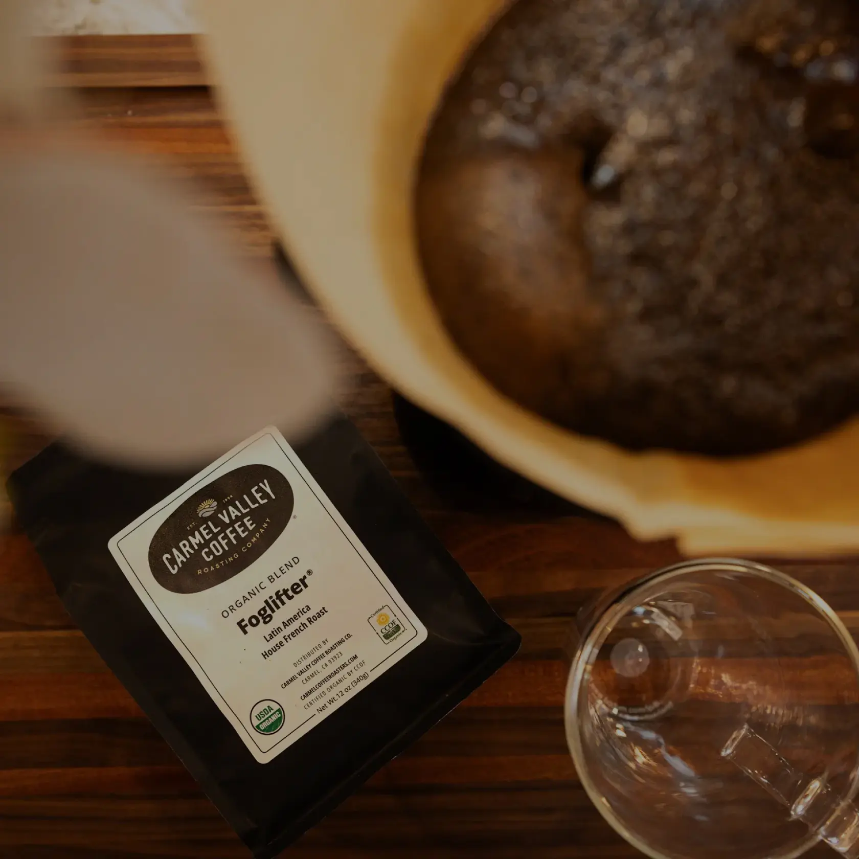 Bag of Carmel Valley Coffee Foglifter blend beside coffee brewing equipment and a glass mug on a wooden surface.