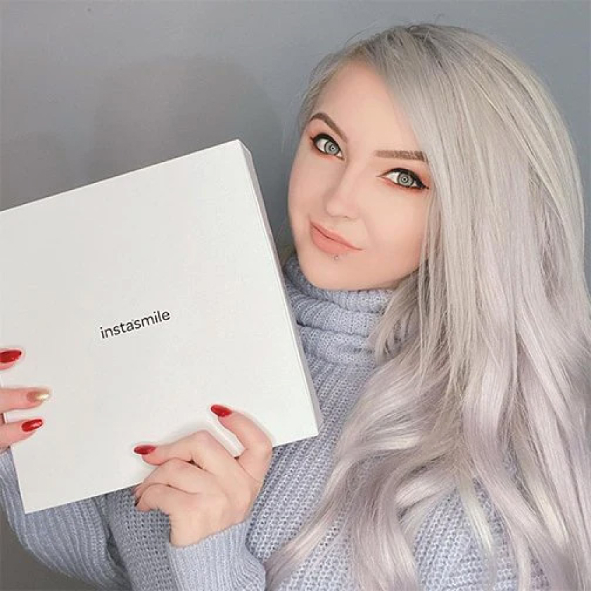 A woman with long, platinum blonde hair and a soft smile holds up a white box labeled “instasmile.” She’s wearing a cozy lavender knit sweater and has neatly manicured red nails. The minimalist background and box design highlight the branding, suggesting a product reveal or customer endorsement for a cosmetic dental solution.