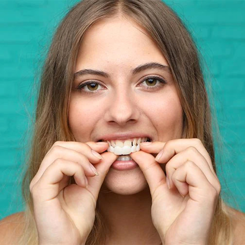 A young woman with long, light brown hair is placing a removable dental veneer over her upper teeth using both hands. She is looking forward with a slight smile during the process. The background is a solid teal color, highlighting the cosmetic dental product and its easy, at-home application.