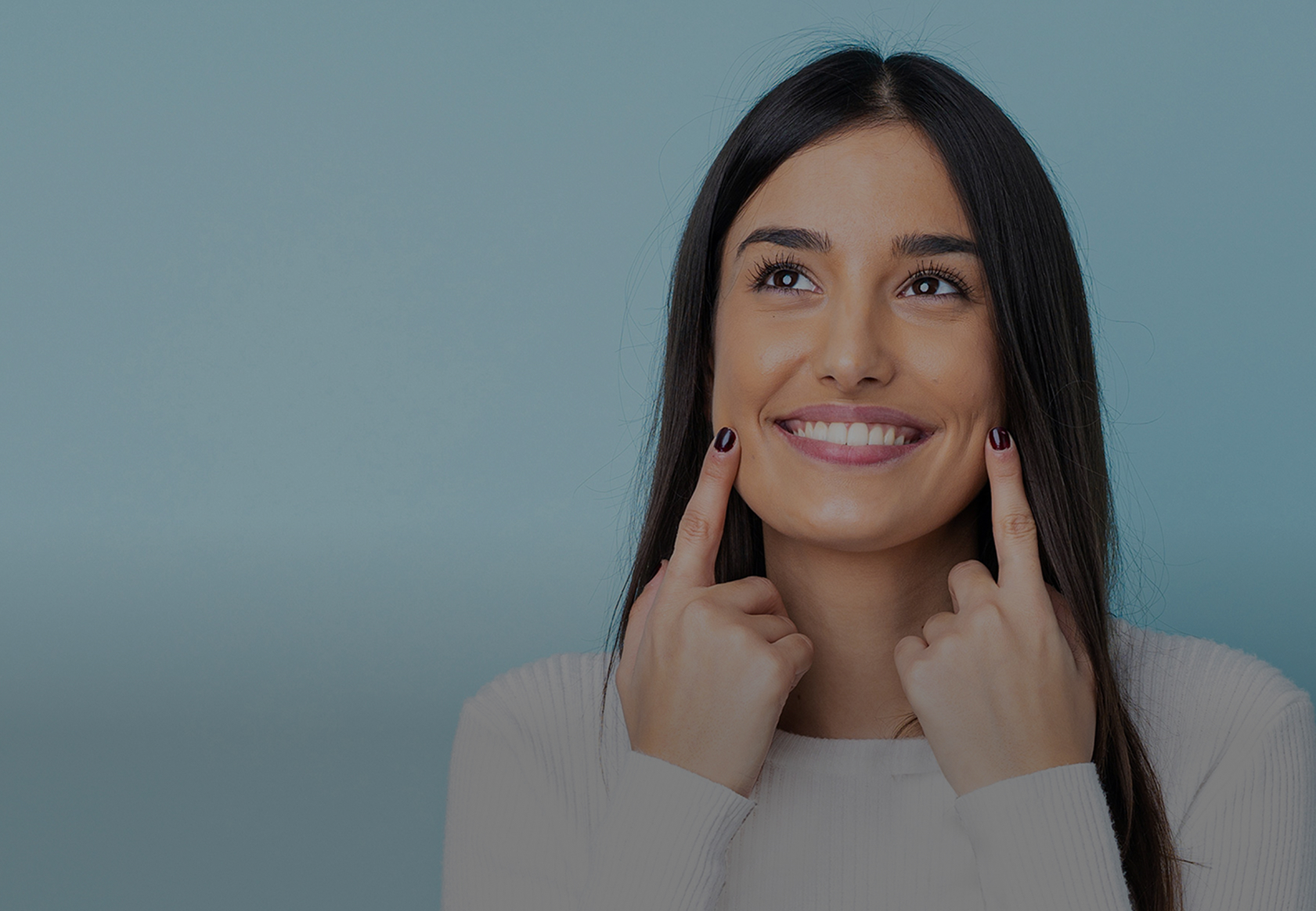A young woman with long dark hair and a white sweater smiles brightly while pointing to her dimples with both index fingers. She looks slightly upward with a joyful expression against a soft blue background, suggesting positivity, self-confidence, or satisfaction—possibly in relation to dental, beauty, or wellness themes.