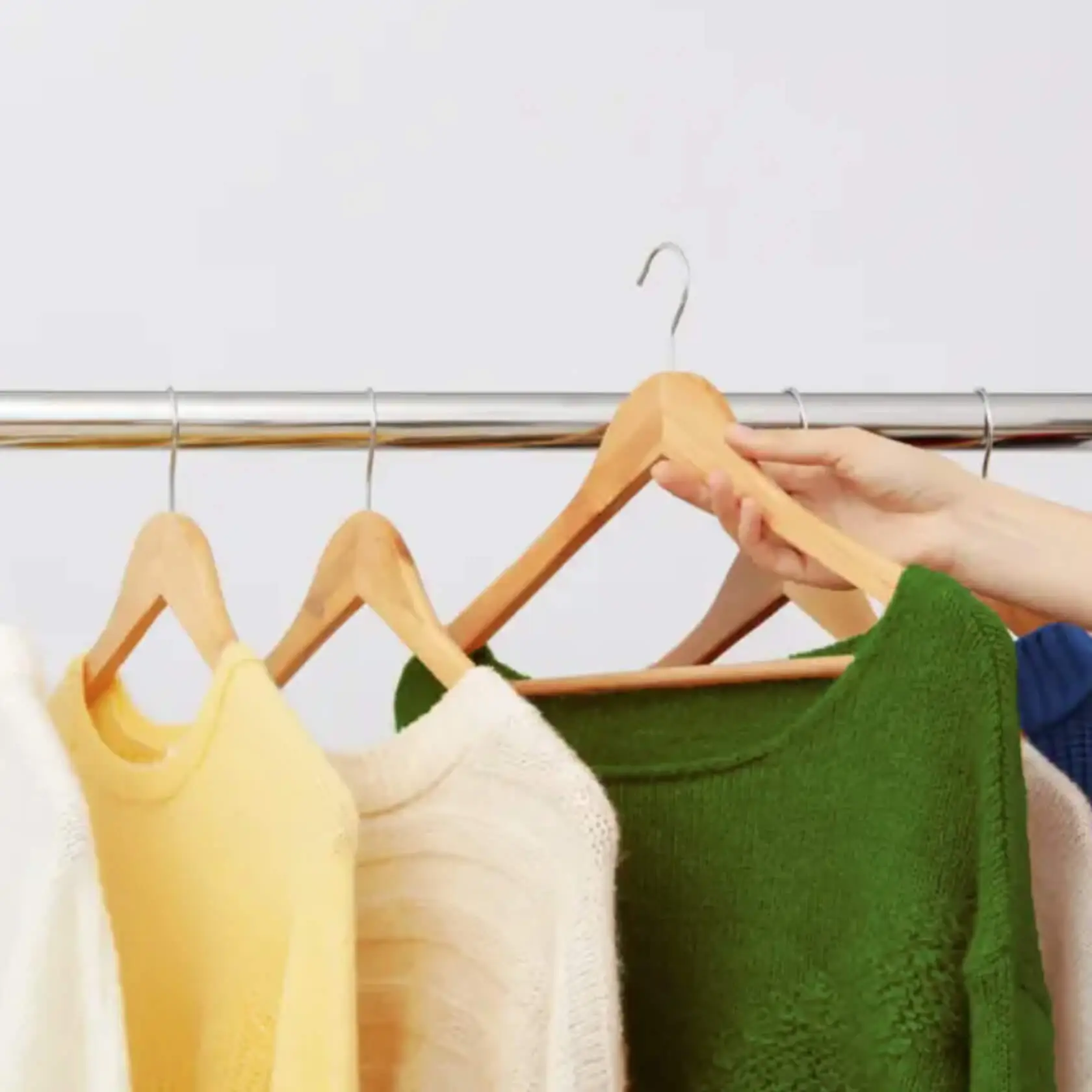 A hand reaches for a green sweater among knit sweaters in white, yellow, and cream hanging on a chrome clothing rail.