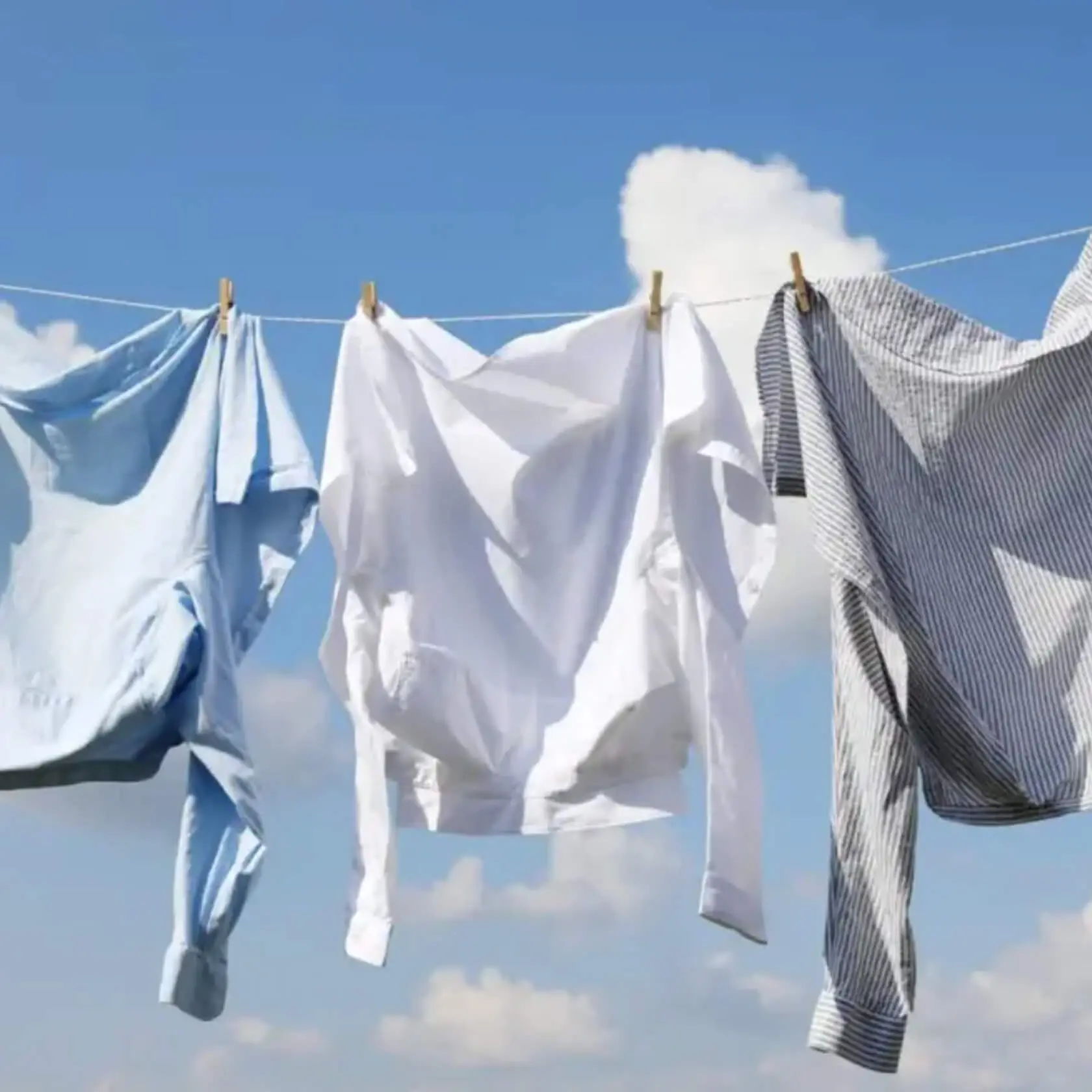 Three shirts—light blue, white, and blue-striped—pinned to a clothesline, drying against a bright blue sky with clouds.