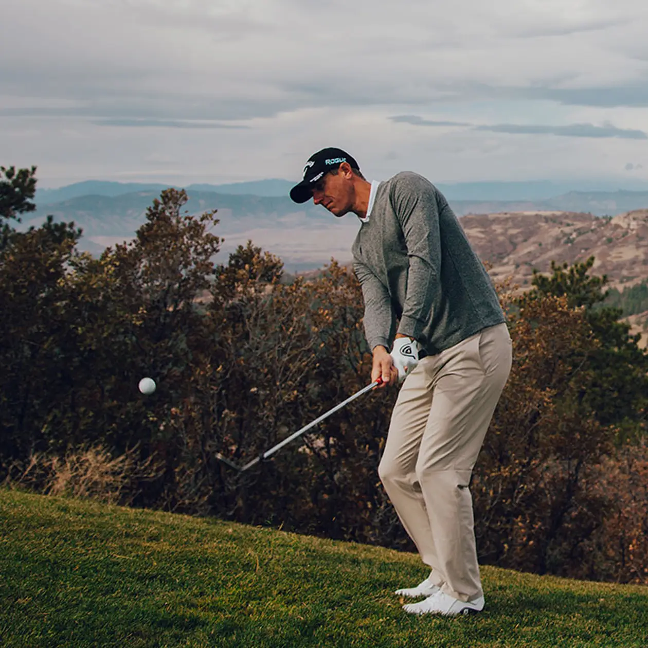 A golfer in a dark gray long-sleeve shirt, beige pants, and a black cap is mid-swing on a grassy slope surrounded by trees and rugged terrain. The background features a scenic mountain range under a cloudy sky, capturing a moment of focus and athletic precision in a natural outdoor setting.