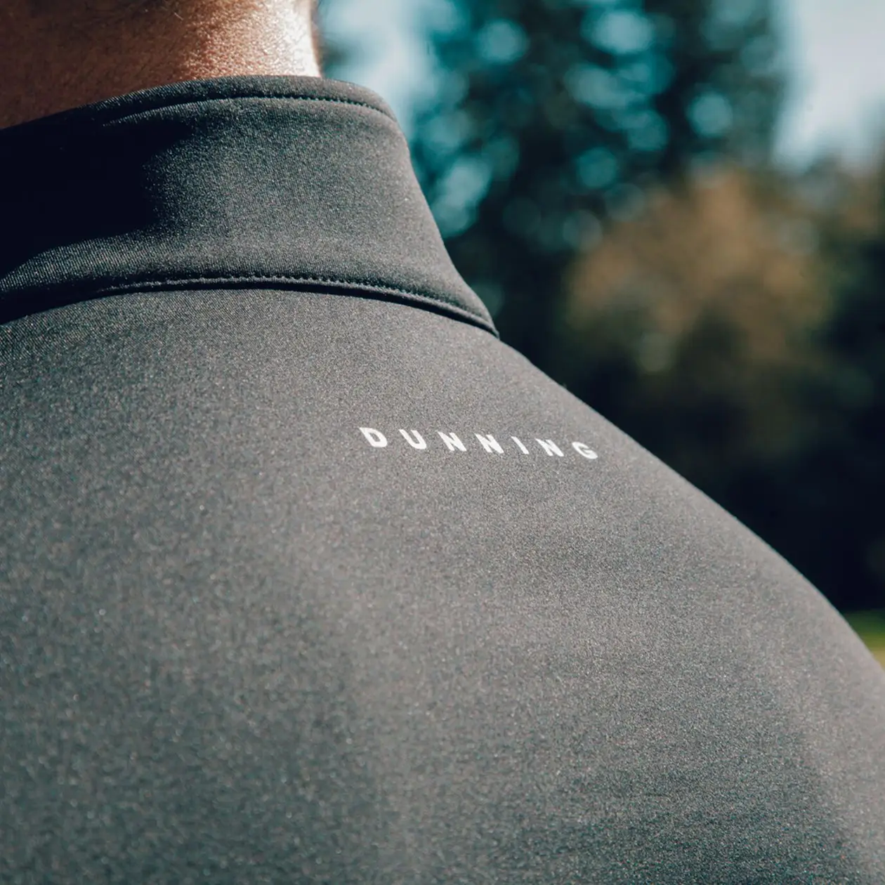 Close-up of the back of a dark athletic garment with the brand name “DUNNING” printed in white capital letters across the upper back. The fabric appears smooth and performance-oriented, with blurred trees and sunlight in the background suggesting an outdoor golf setting.