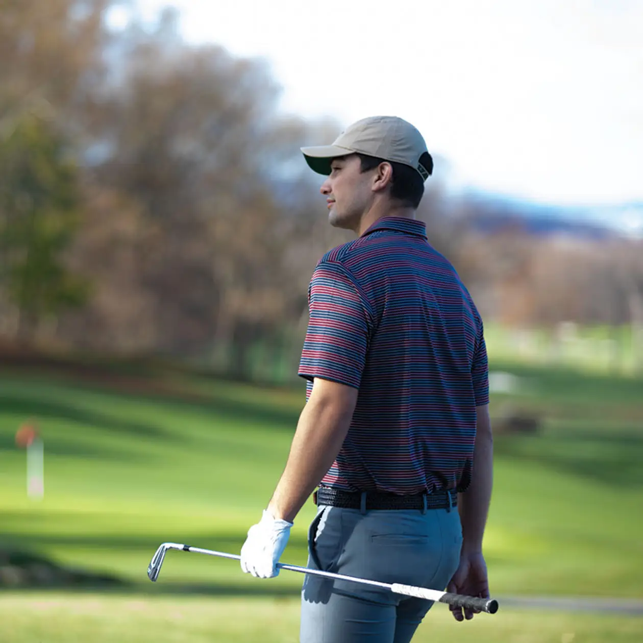 A man in a striped golf shirt, gray pants, and a beige cap stands on a green golf course, holding a club in one hand and looking into the distance. The background features soft-focus trees and rolling fairways, suggesting a peaceful and reflective moment during a round of golf.