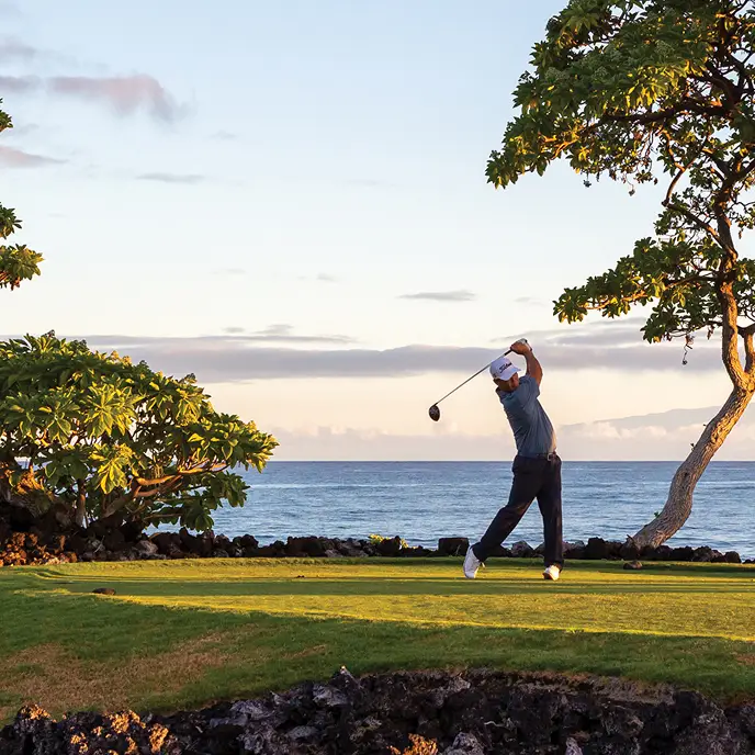 A golfer swings a club on a scenic coastal course surrounded by dramatic lava rock, lush green grass, and uniquely shaped tropical trees. The ocean stretches out in the background under a pastel-colored sky at sunset, capturing a serene and picturesque moment of outdoor leisure.