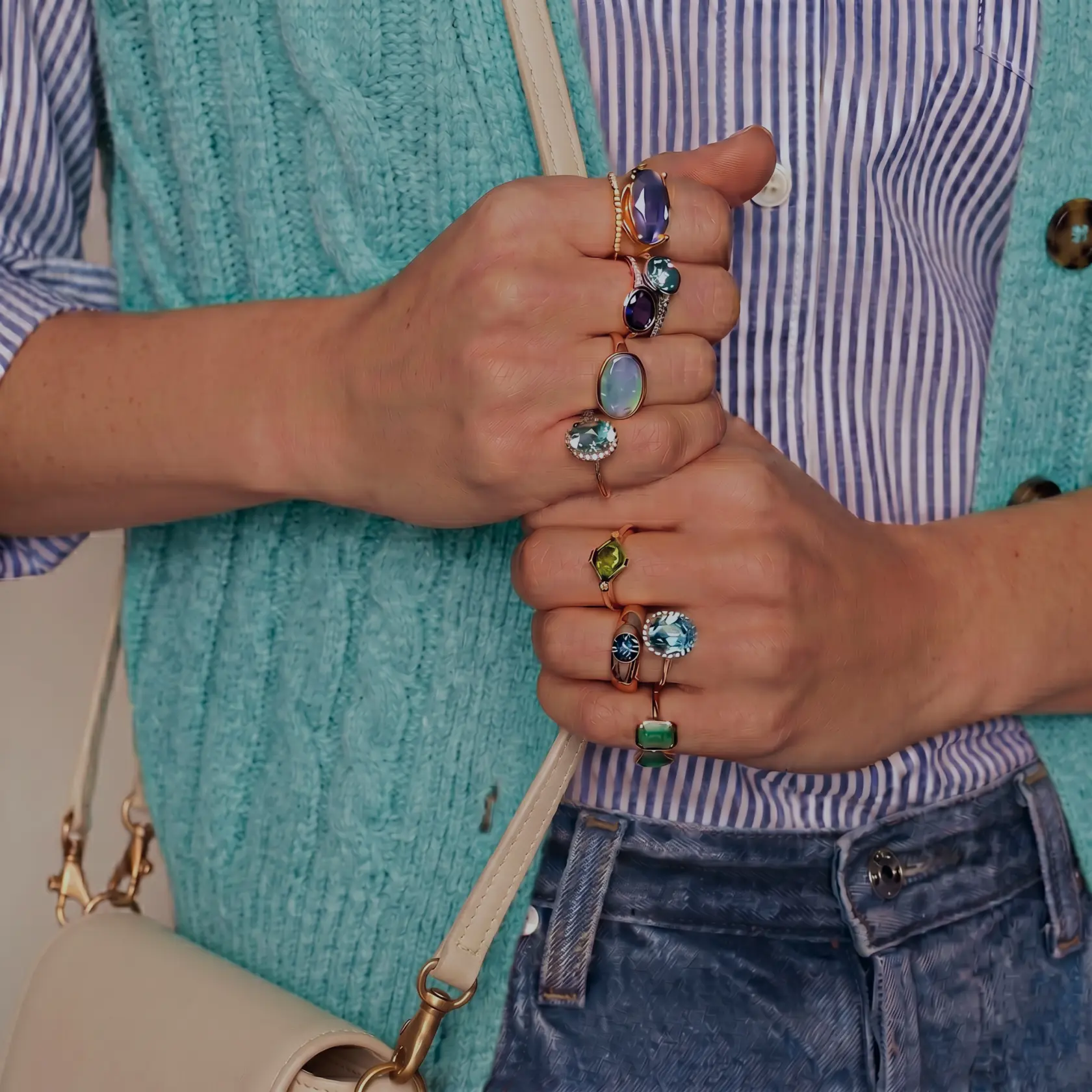 Close-up of hands wearing colorful gemstone rings, styled with a striped shirt, turquoise knit vest, jeans, and a cream shoulder bag.