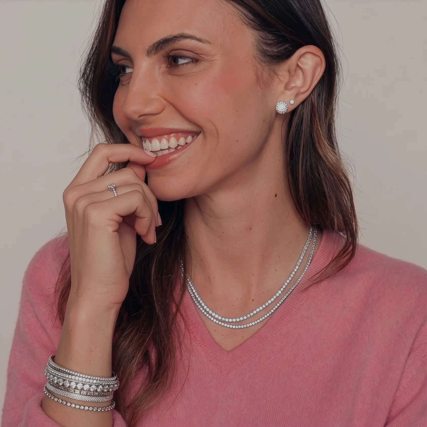 Smiling model wearing layered diamond jewelry, including stud earrings, necklaces, bracelets, and a ring, styled with a pink sweater.